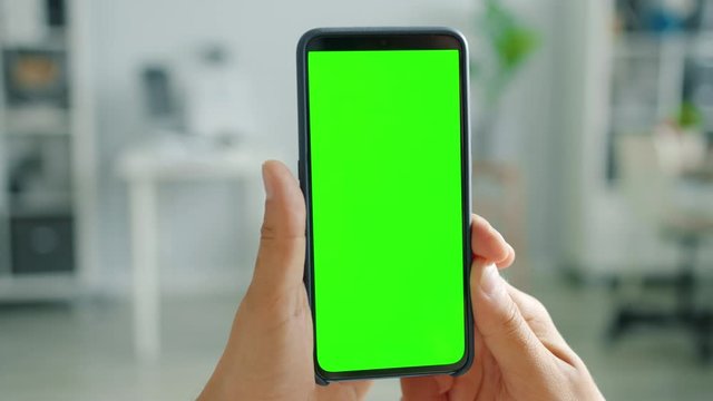 Contemporary smartphone with green screen copy space mock-up in male hand on blurred domestic background. Digital devices and online content concept.