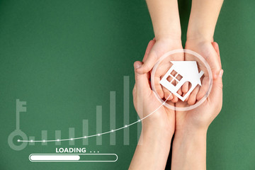 Loading with female and child hands holding white paper house on green background. Family home and...