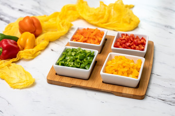 Red, yellow, green, and orange sweet bell peppers chopped in small white dishes on a wooden board; whole peppers in background on yellow material; white marble countertop