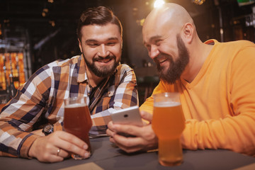 Two friends laughing, using smart phone at beer pub. Excited man showing something online to his friend while drinking together