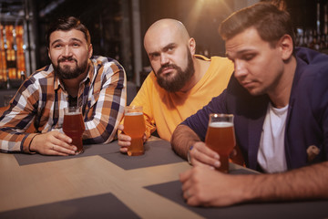 Group of male friends looking upset, drinking beer after watching football game. Three men unhappy after the loss of their favourite team, drinking at beer pub