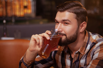 Fototapeta na wymiar Close up of a happy relaxed handsome man sipping delicious beer, looking away cheerfully. Attractive bearded man enjoying glass of beer at the pub