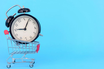 Shopping cart and classic alarm clock on blue background. Sale time buy mall market shop consumer concept. Selective focus.