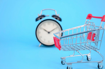 Shopping cart and classic alarm clock on blue background. Sale time buy mall market shop consumer...