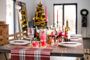 Table served for Christmas dinner with decorations, candles, fruits, glasses and lanterns. Bright living room decorated with lights and Christmas tree. Natural light shot.