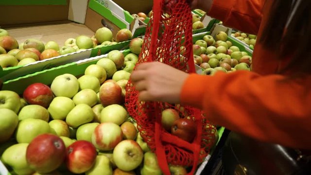 Young woman picking veggies and fruits in supernarket in mesh organic shopping bag, zero waste