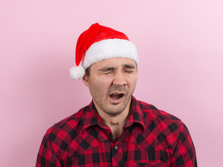 Emotions on the face, tired, holiday hangover, awareness. A man in a plaid rabbit and a Christmas red hat, on a pink background, copy space.