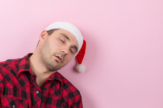 Emotions On The Face, Tired, Holiday Hangover, Awareness. A Man In A Plaid Rabbit And A Christmas Red Hat, On A Pink Background, Copy Space.