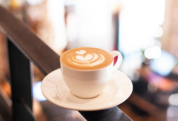 Cup of cappuccino with latte art stands on the background of the cafe in defocus. White ceramic cup and place for text.