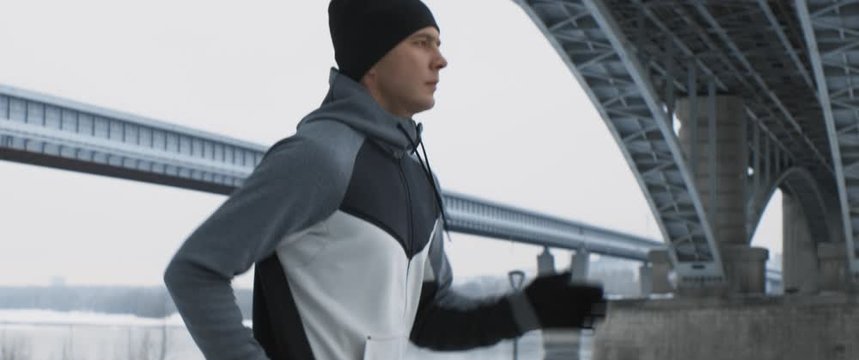 Young male caucasian strong runner jogs along the embankment of big city in cold winter. Urban environment, bridge, promenade in siberia. Anamorphic portrait shot