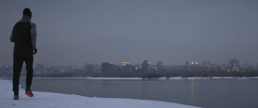 Unrecognizable male athlete completes winter run in snow on river bank in dusk. Jogging, outdoor sport, city lights, cold season, industrial urban environment. Anamorphic shot