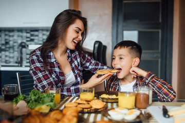 Cheerful brunette mother feeding her son sandwich with cheese during breakfast at home.