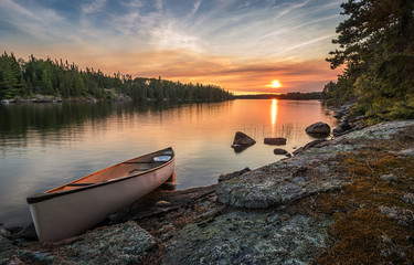 A lone canoe on shore at a peaceful lake at sunset. The odd colored clouds in the background are from a forest fire in Northwest Ontario, Canada in the summer of 2018. - obrazy, fototapety, plakaty
