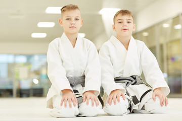 Portrait of two little boys in white kimono sitting on the floor and looking at camera they doing karate in gym