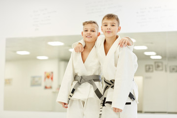 Portrait of two boys in kimono standing and embracing each other and smiling at camera after karate...