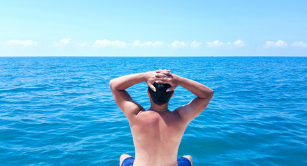 Rear view of young man with naked torso sitting on the edge of yacht looking at seascape.