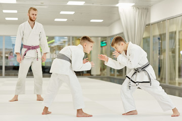 Two boys in kimono learning to fight during training in karate while their coach standing and...