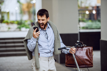 Nervous bearded fashionable caucasian employee pushing his bike, holding smart phone and yelling....