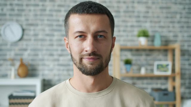 Slow motion portrait of young man serious then smiling in cozy house looking at camera standing alone. People, positive emotions and lifestyle concept.