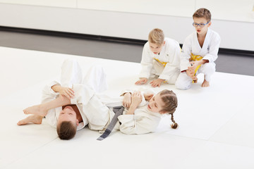Girl fighting with boy on the floor while other children cheering for them during lesson in karate...
