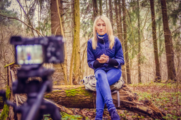 blonde girl in a blue jacket and jeans, boots writes a blog like a blogger sitting in a forest, Park on a fallen tree. Writes a blog on the DSLR.