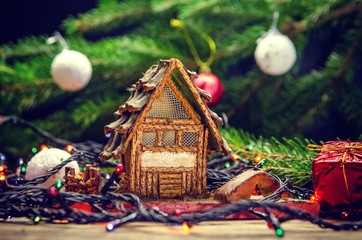 Toy house among the Christmas lights, small red gifts on the background of the branches of the Christmas tree with balls close-up. Beautiful Christmas and New year background. Tinted photo