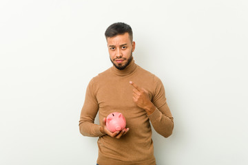 Young south-asian man holding a piggy bank pointing with finger at you as if inviting come closer.