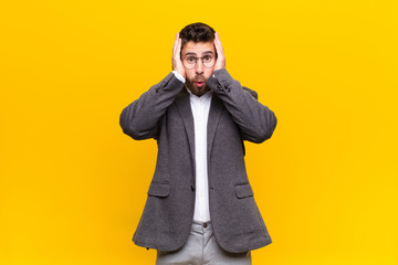 young handosme man looking unpleasantly shocked, scared or worried, mouth wide open and covering both ears with hands against flat color wall