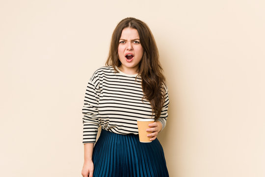 Young curvy woman holding a coffee screaming very angry and aggressive.