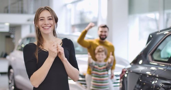 Cheerful Caucasian girl bragging car keys in showroom. Adult man and little boy showing thumbs up at the background. Successful businesslady buying new vehicle for family. Cinema 4k footage ProRes HQ.