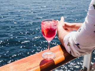 Fashionable man holding a beautiful glass of pink wine on the open deck of a cruise liner against the backdrop of blue sea waves. Side view, close-up. Concept of leisure and travel