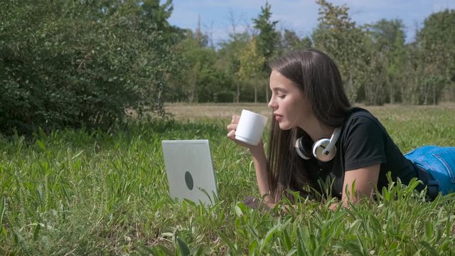In grass with a gadget. Cute girl on her hand with a laptop and a cup of coffee.