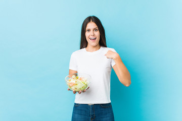 Young caucasian woman holding a salad surprised pointing at himself, smiling broadly.