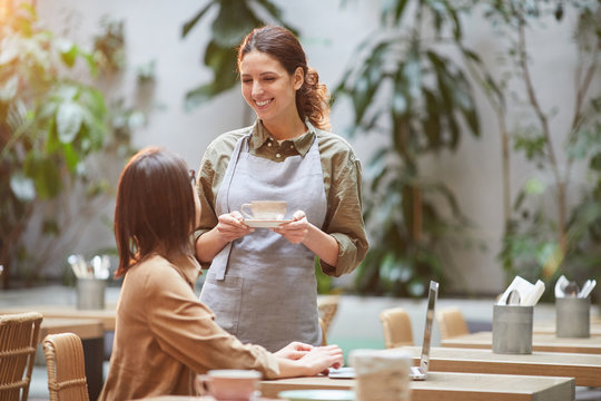 Portrait of smiling young waitress bringing coffee to client at outdoor cafe terrace lit by warm sunlight, copy space