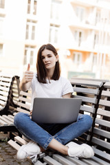Young beautiful woman with thumbs up working on a laptop sitting on bench in the street