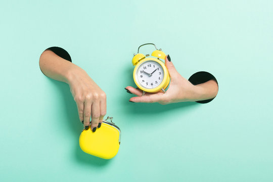 Female hands hold alarm clock and wallet through a hole on neon mint background. Time is money concept.