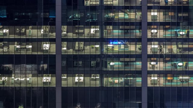 Office building exterior during late evening with interior lights on and people working inside night timelapse. Aerial close up view from above with many illuminated windows. Zoom in