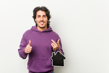 Young man holding a house icon shape smiling and raising thumb up