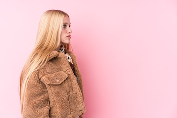 Young blonde woman wearing a coat against a pink background gazing left, sideways pose.