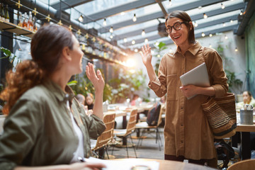 Portrait of two young women waving at each other during meeting in cafe on outdoor terrace...