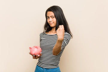 Young asian woman holding a piggy bank showing fist to camera, aggressive facial expression.