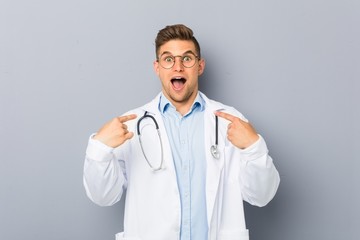 Young blonde doctor man surprised pointing at himself, smiling broadly.