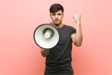 Young hispanic man holding a megaphone showing fist to camera, aggressive facial expression.