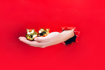 Female hand holding two golden Christmas tree baubles through a hole in red background.