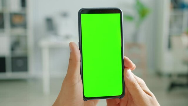 Green screen chroma key smartphone is held my male hands indoors in apartment, person is watching content. Modern technology and information concept.
