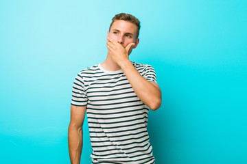 Young caucasian man against a blue wall thoughtful looking to a copy space covering mouth with hand.