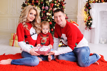 young family-mom, dad and little son in Christmas sweaters with reindeer on the background of the Christmas tree. Parents give a Christmas gift to their child.