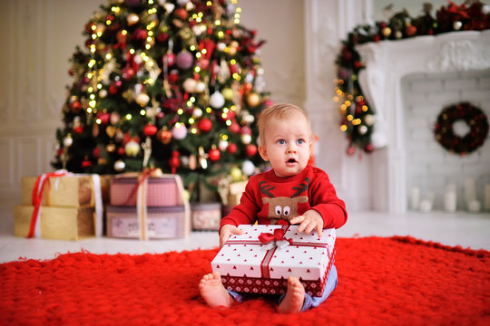 a child-a little boy in a red Christmas sweater with a picture of Santa's reindeer sits barefoot on a warm red carpet with a gift in his hands on the background of a Christmas tree and decor