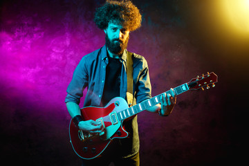 Obraz premium Young hipster man with curly hair with red guitar in neon lights. Rock musician is playing electrical guitar.
