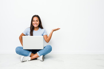 Young mixed race indian woman sitting working on laptop showing a copy space on a palm and holding another hand on waist.